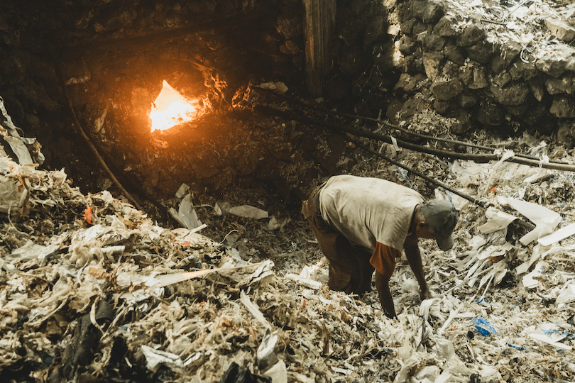 A person bends over, sorting through piles of trash near a large open fire in an industrial setting. The stone walls and scattered debris indicate a challenging work environment.