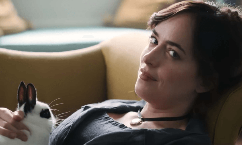 Actor Dakota Johnson lying on a couch with a black and white rabbit on her lap. She is looking at the camera.