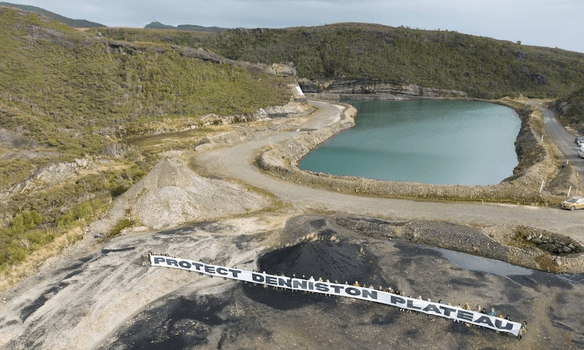 a lake and a shallow pit of dark earth surrounded by bouncy green bush, with a line of people holding a banner that reads "protect denniston plateau"