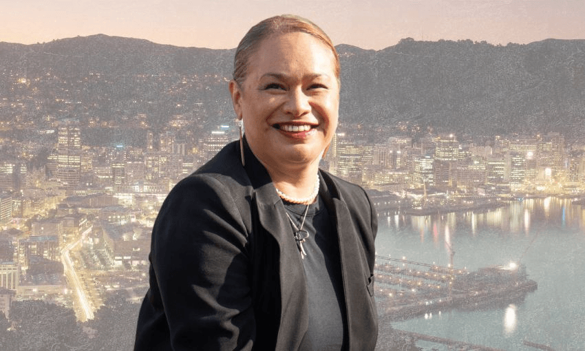 A woman in a black blazer smiles at the camera with a cityscape and harbor in the background during dusk.