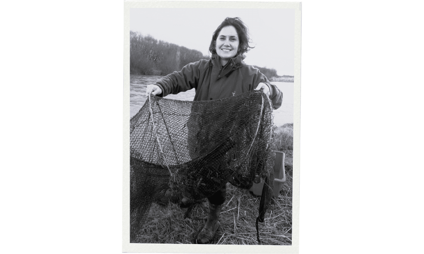 A smiling researcher, Jane Kitson, stands outdoors by a river, holding up a large fishing net. The scene appears to be during colder weather, as they are wearing a jacket. The image is in black and white.
