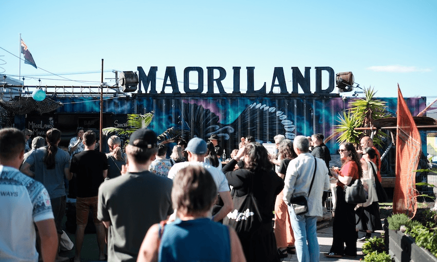 A crowd of people walk towards a large sign reading "Māoriland"