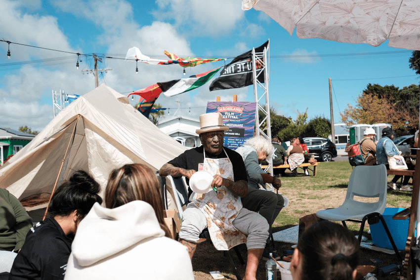 Activist Tame Iti sits outside at Māoriland festival with a palestine and toitū te tiriti flag flying in the background