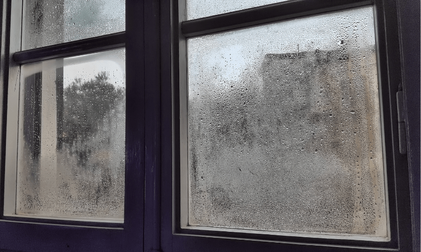 A close-up view of a window with condensation and raindrops on the glass, blurring the view of trees and a building outside. The frame is dark-colored, creating a contrast with the lighter outdoor scene.