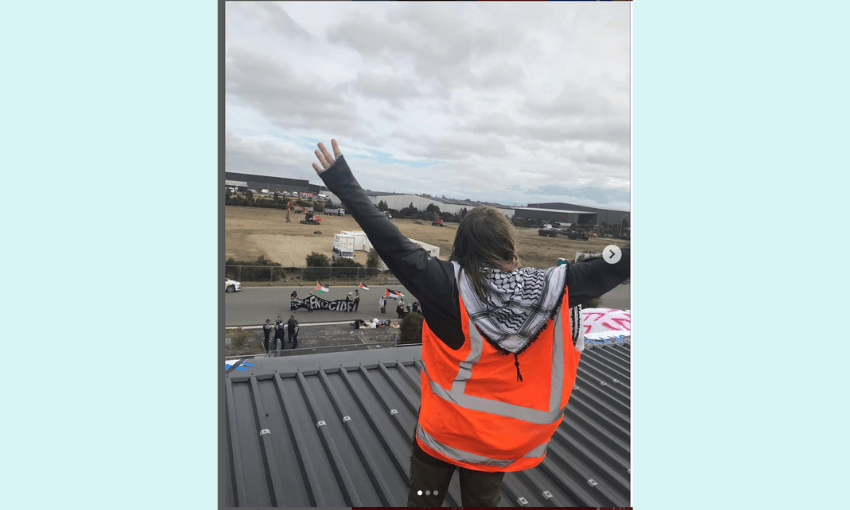 an image of a person on a roof with a pale blue background, from behind with their arms up and wearing a black and white palestinian kuffiyah