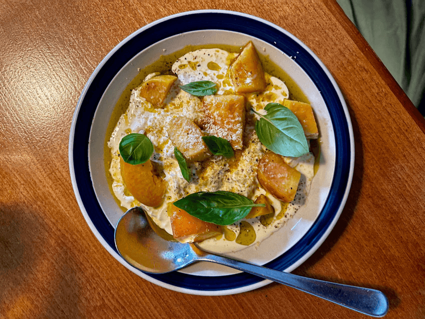 A bowl of creamy stracciatella cheese topped with pieces of peach, basil leaves, and grated cheese. A silver spoon rests on the bowl's edge, which is on a wooden table. Drizzles of olive oil and pepper are visible.