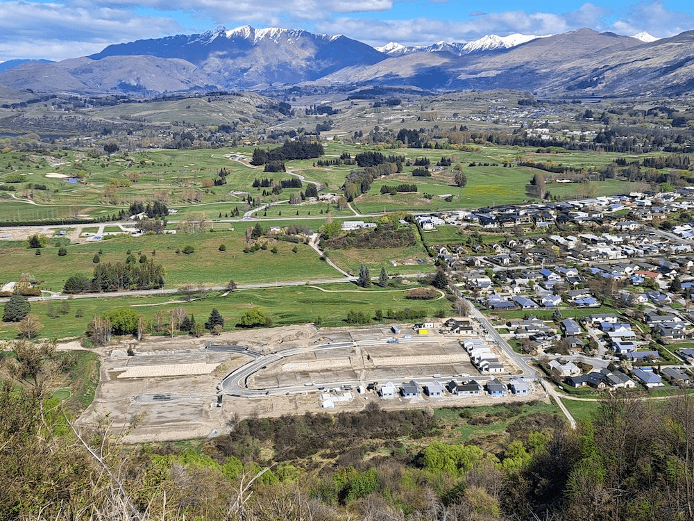 a green field with a big emty dirty lot that has some foundations laid in it. Blue skies, mountains in the background and bright green paddocks