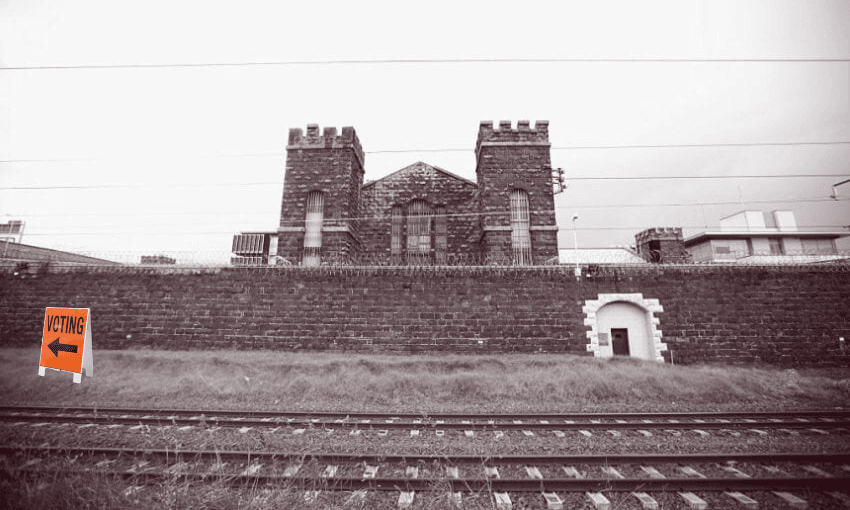 A large stone prison building stands behind a high wall and railway tracks. A bright orange sign on the grass reads "VOTING" with an arrow pointing left.