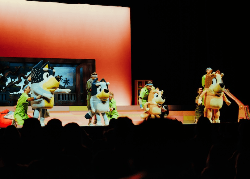 People in green outfits control large dog character costumes on stage in front of an orange-lit backdrop, performing for an audience in a theatre setting.