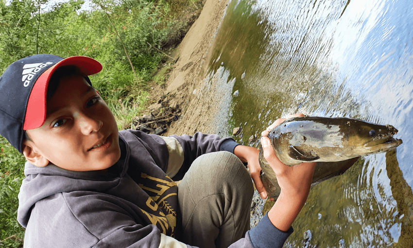 A boy wearing a red and black cap and grey hoodie kneels by a riverside, smiling as he holds a large tuna (freshwater eel) with both hands near the water’s edge. The background shows green foliage and a sandy bank.