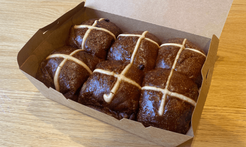 An open cardboard box on a wooden table, containing six dark brown hot cross buns with white icing crosses on top