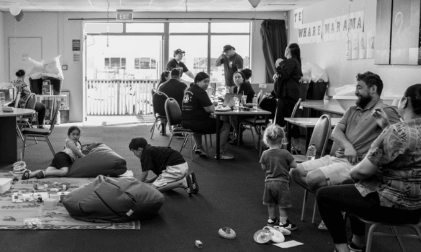 Black and white image of a community center. Adults sit at tables, some chatting. Children play on the floor with toys and bean bags. Sunlight streams through large windows in the background. A sign on the wall reads "Te Whare Marama.