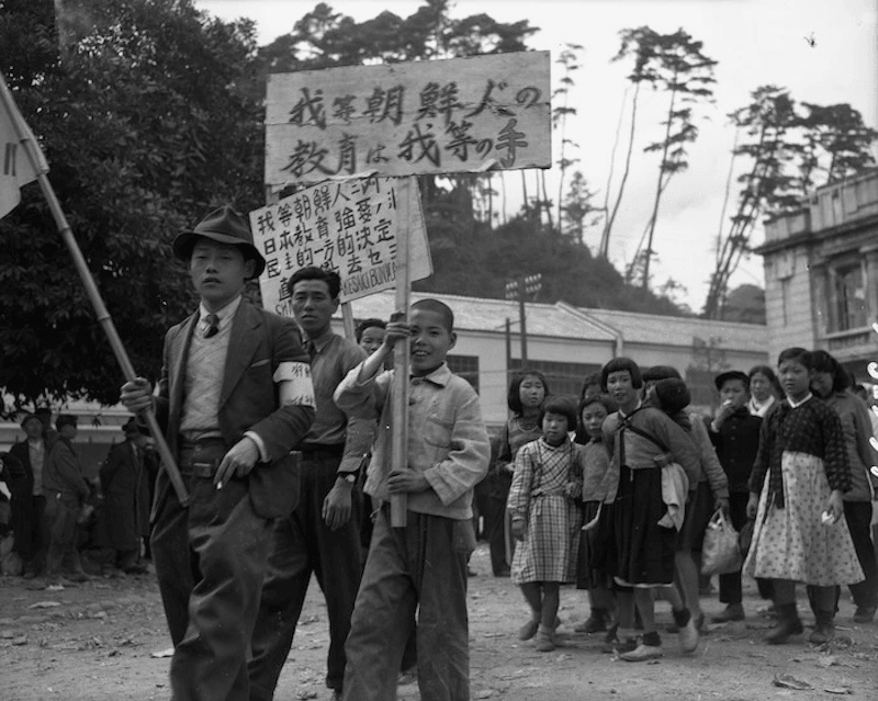 A group of children and adults march outdoors, carrying banners with Japanese text. The crowd is dressed in mid-20th-century clothing, and trees and a building appear in the background.