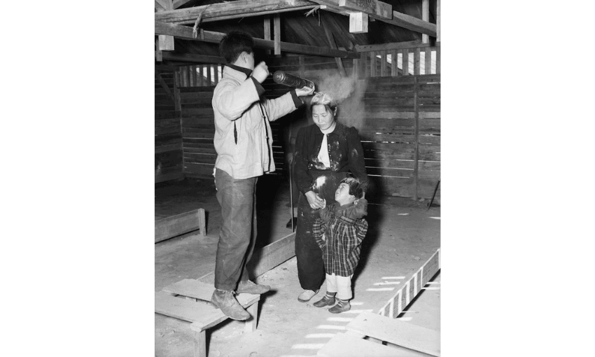 A young man stands on a bench and sprays liquid onto a woman’s head while a small child hugs her. They are inside a wooden structure with exposed beams and slatted walls.