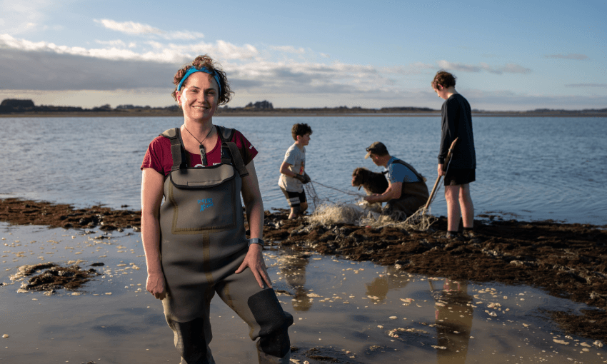 Jane Kitson in waders stands smiling by the shore, while three people and a dog work with nets in shallow water behind her under a blue sky.