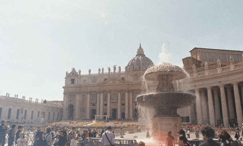 Crowds gather around a large ornate fountain in St. Peter's Square, Vatican City, with St. Peter's Basilica in the background under a bright, sunny sky.
