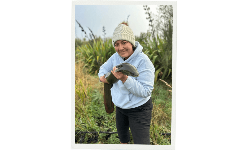 Erina Watene, in a white knit hat and light hoodie smiles while holding a large tuna or freshwater eel, surrounded by tall harakeke fronds and leafy plants.