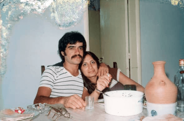 A man with a mustache and a woman sit closely together at a table, smiling slightly. The table has dishes, a ceramic jug, bottles, and utensils. The photo appears old and has some damage at the edges.