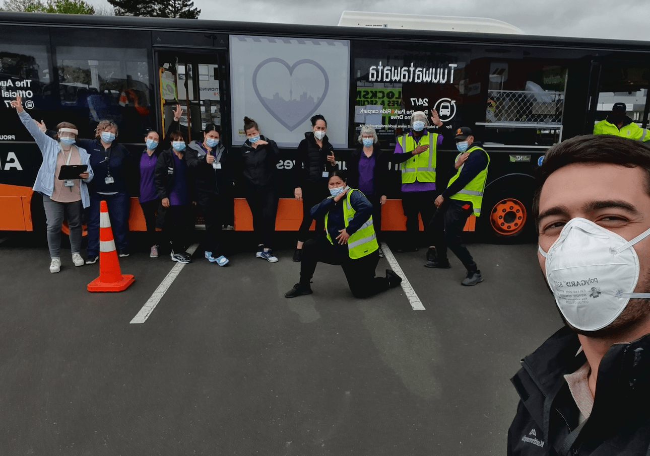 A man wearing a face masks takes a selfie in front of a vaccination bus and a group of people wearing masks and hi-vis