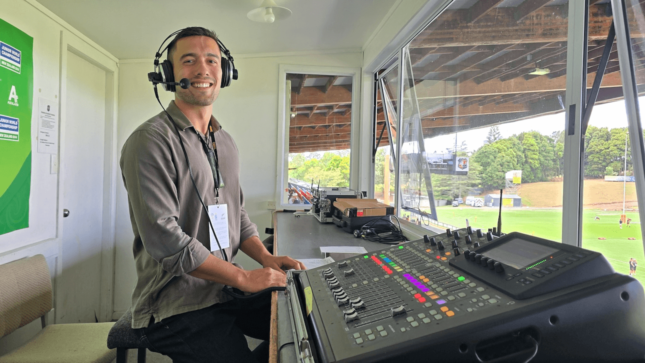 A man in a polo shirt wears a headset and stands in front of a control desk in a commentary box