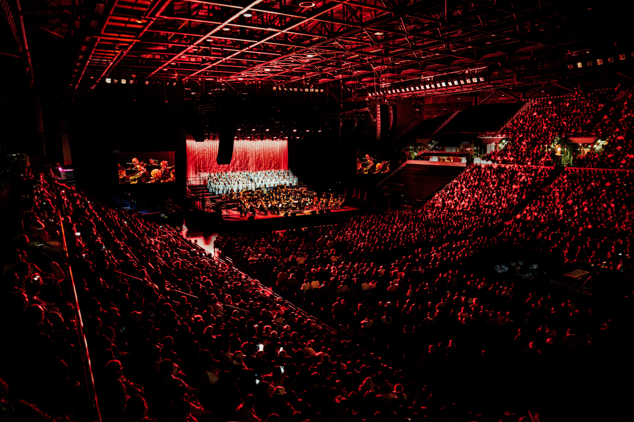 A large audience fills an indoor arena, all facing a brightly lit stage where an orchestra performs under red lighting, creating a dramatic atmosphere. The venue is packed and the mood is vibrant.