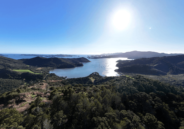 A scenic view of a bay surrounded by forested hills, with small islands in the distance and the sun shining brightly over a calm, blue sea.
