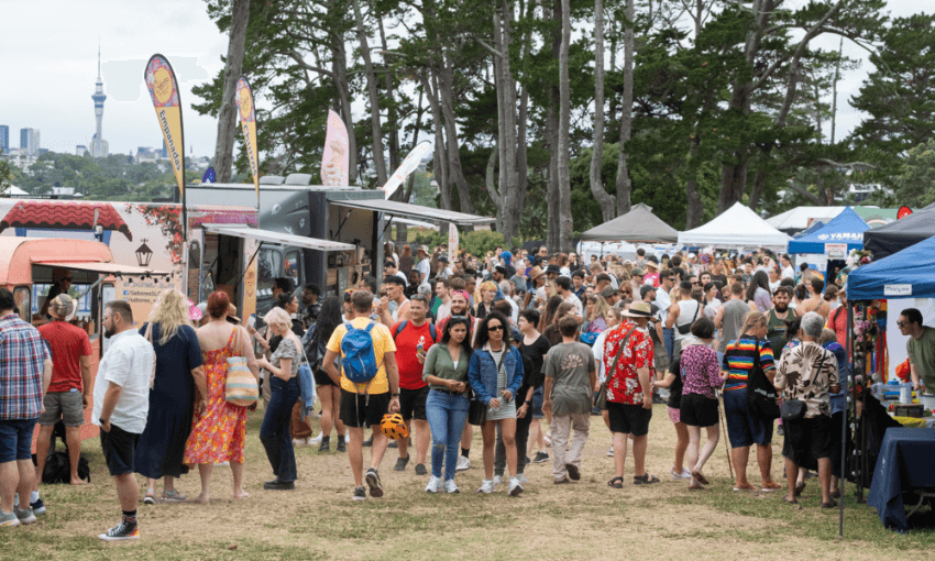 People in shorts and t-shirts wander through a group of stalls selling food at a summer festival. The Auckland sky tower is visible in the background. The event looks laid back and fun.