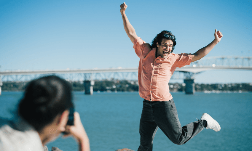 A man in a short sleeve button down shirt and jeans is jumping in the air in front of a woman who is taking a picture of him, the Auckland Harbour bridge and the sparkly, aqua harbour is behind him.