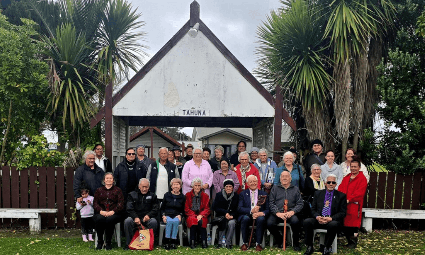 A large group of people, including elders and children, pose for a photo outside a traditional building with a sign reading "TĀHUNA." Tī kouka cabbage trees and greenery surround the scene, and everyone is dressed warmly.