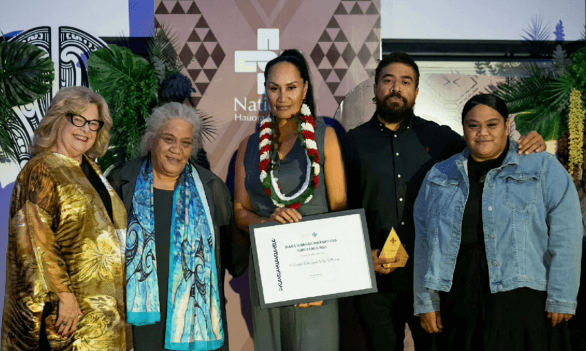 Five people stand together at an award ceremony; the central person holds a certificate and wears a floral lei. The group is dressed in formal attire, smiling in front of a decorated backdrop with patterns and greenery.