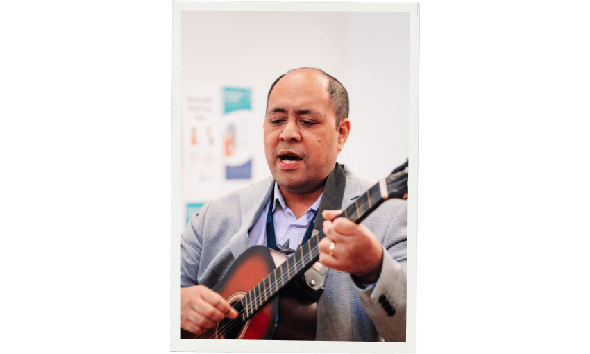 A man in a gray jacket and blue button up shirt plays guitar and sings in a light filled environment that looks like a classroom.