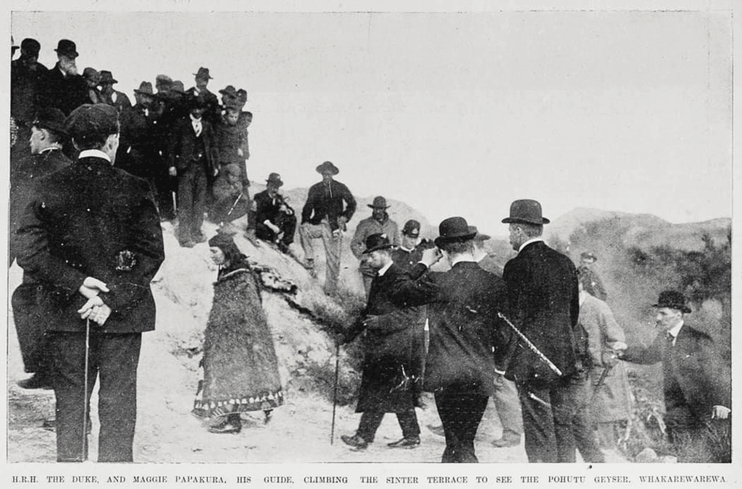 A group of men in formal clothing, some with canes and hats, stand and watch as a woman in traditional dress guides another man up a rocky hillside. Text below identifies the guide as Maggie Papakura.
