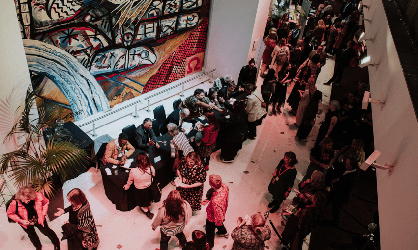 An overhead shot of a crowd lining up in a large foyer with a large scale painting on the walls. There are people seated at a table - they are signing books.