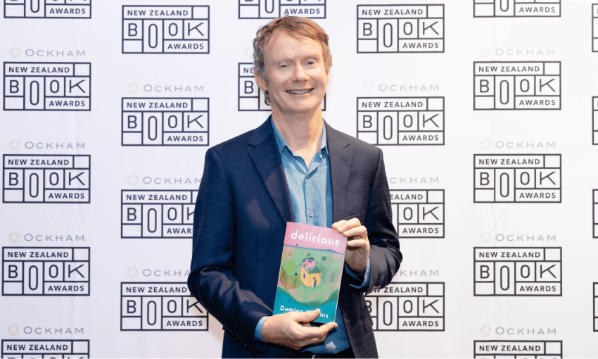 A photograph of author Damien Wilkins holding his novel and standing against a background of Ockham logos.