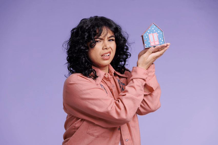 A person with curly hair in a pink shirt holds a small model house and looks confused or skeptical, standing against a purple background.