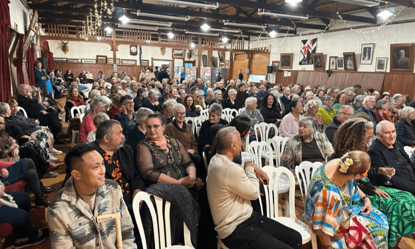 A photograph of a crowd of people seated on white chairs in an old fashioned village hall.