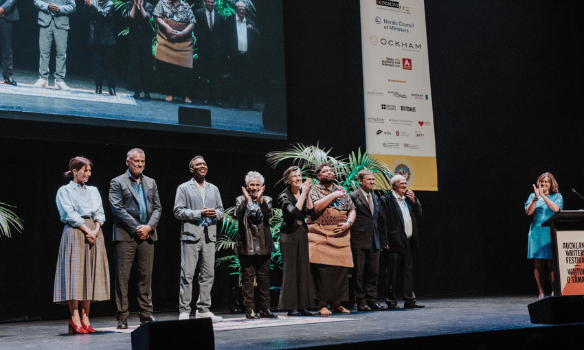 A photograph of adults standing in a line on a large stage with a big screen behind them. 