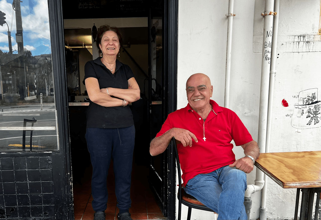 A woman stands smiling with arms crossed in a doorway, while a man in a red shirt sits on a stool next to her outside, also smiling. They appear relaxed and happy.
