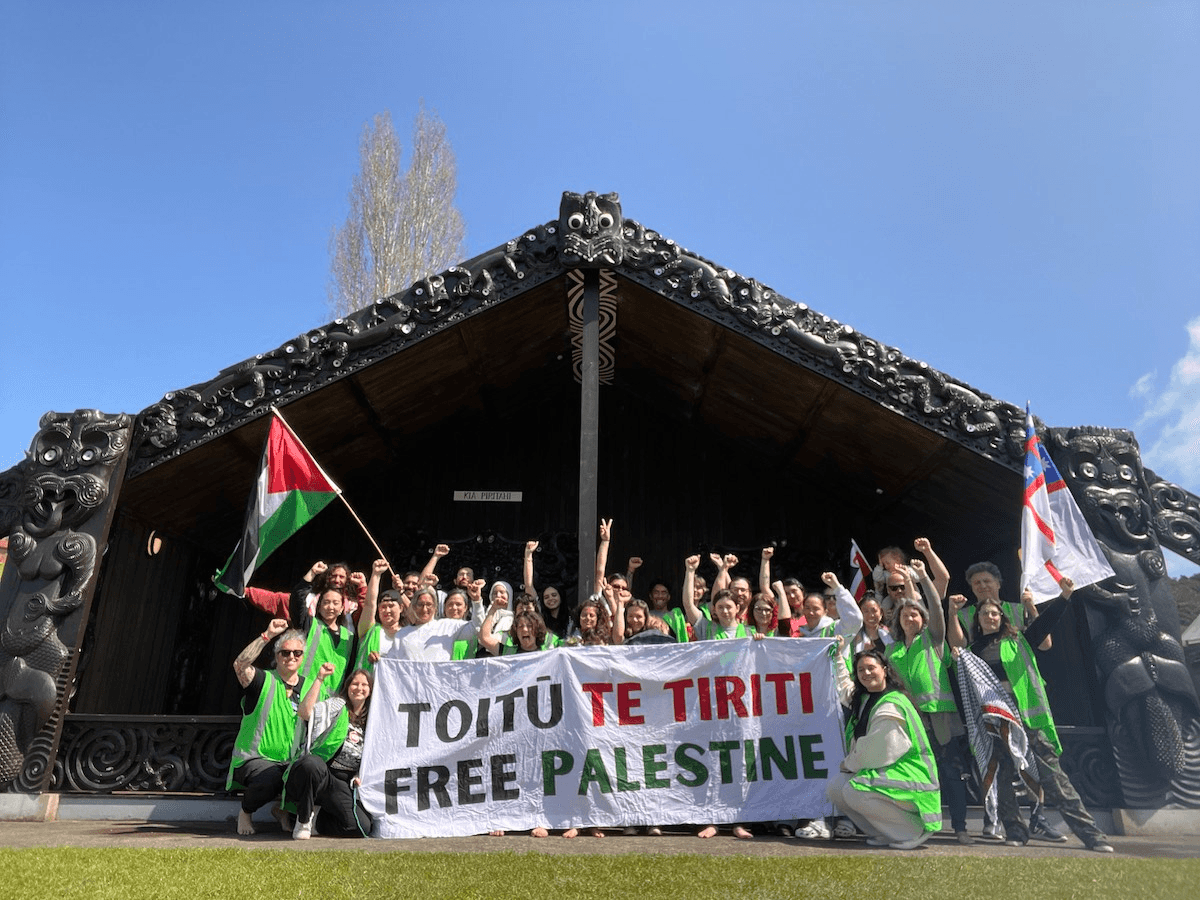 people in fluro vests standing front of a marae holding a banner that reads "Toitu te tiriti, Free Palestine"