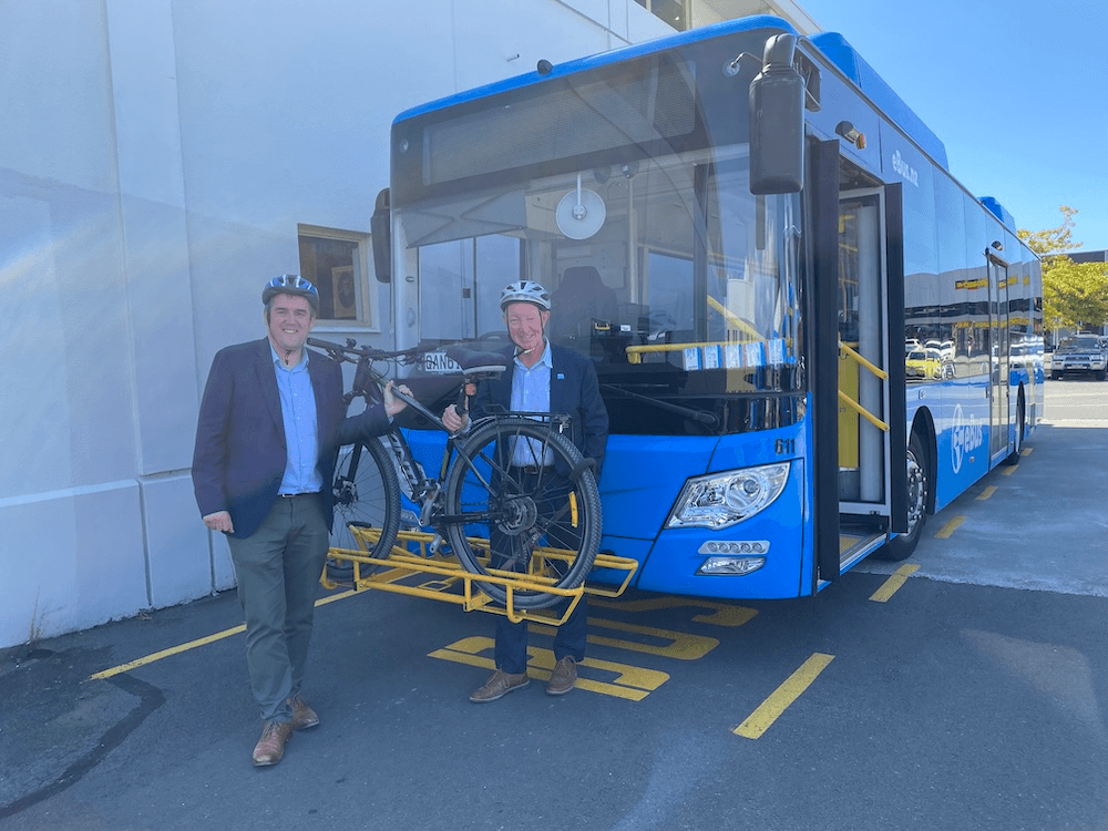 a bus on a sunny day, two men in business casual with blue shirts and blazers stand next to the bus wearing helments and looking at a bike mounted on a rack at the front