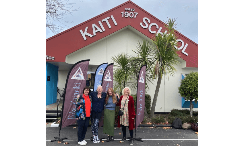 Photo of four women standing in front of a school building with the words Kaiti School painted on the roof facade. 