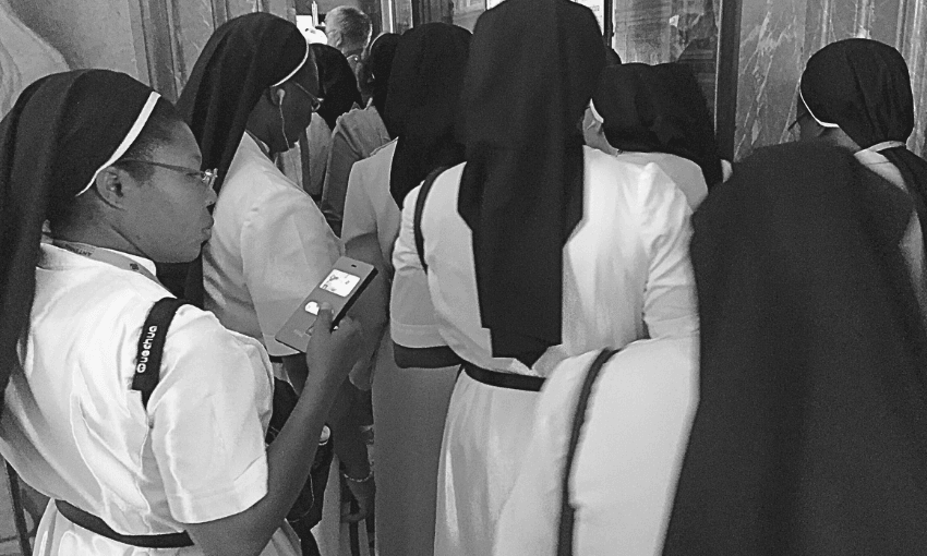 A group of nuns, dressed in white habits with black veils, stand closely together. One nun on the left looks at her phone while the others face away, appearing to wait or move forward in a crowded hallway.