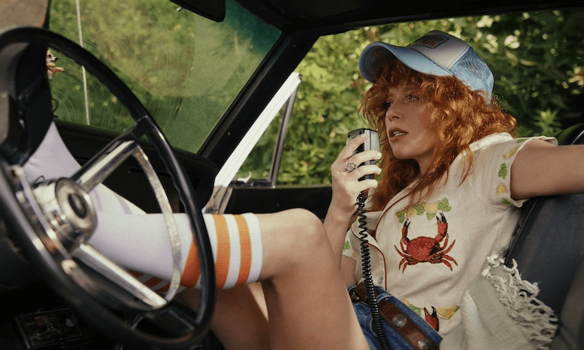 A woman with red curly hair sits inside a car, with her feet on the steering wheel. She is talking into a hand-held radio receiver and wearing a blue cap. She has knee-high white socks with an orange stripe on them.