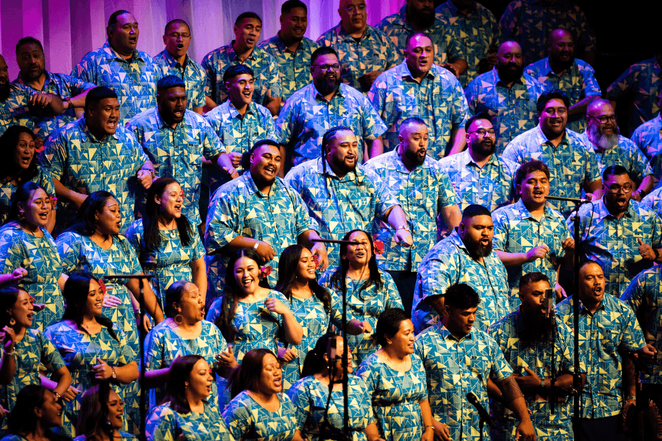 A large choir dressed in matching blue and green patterned outfits joyfully sings on stage under colourful lighting.