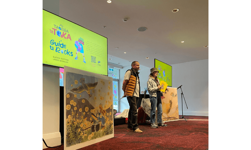 A photograph of two people standing on carpet between two screens showing illustrations from their picture book A Guide to Rocks. 