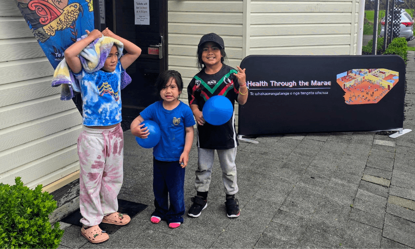 Three children stand smiling outside a building. One holds a blue balloon, another has a towel on their head, and a third wears a cap. A sign nearby reads, "Health Through the Marae."