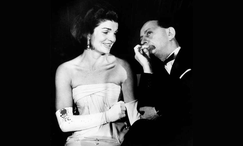 A woman in a strapless dress and long gloves (Jacqueline Kennedy) smiles while sitting beside a man (André Malraux) in a tuxedo, who is resting his chin on his hand and appears to be speaking to her. The image is in black and white.