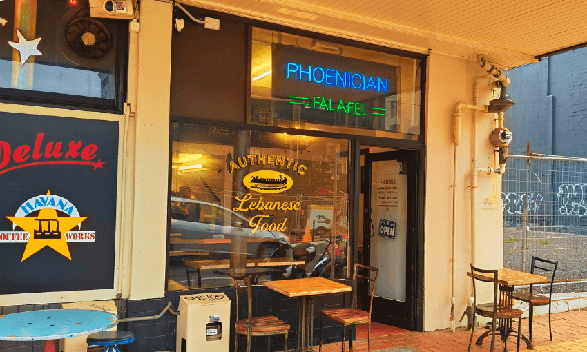 Street view of a small restaurant with signs reading "PHOENICIAN FALAFEL" in neon and "AUTHENTIC Lebanese Food" on the window. Outdoor tables and chairs line the brick sidewalk. A "Havana Coffee Works" sign is visible on the left.
