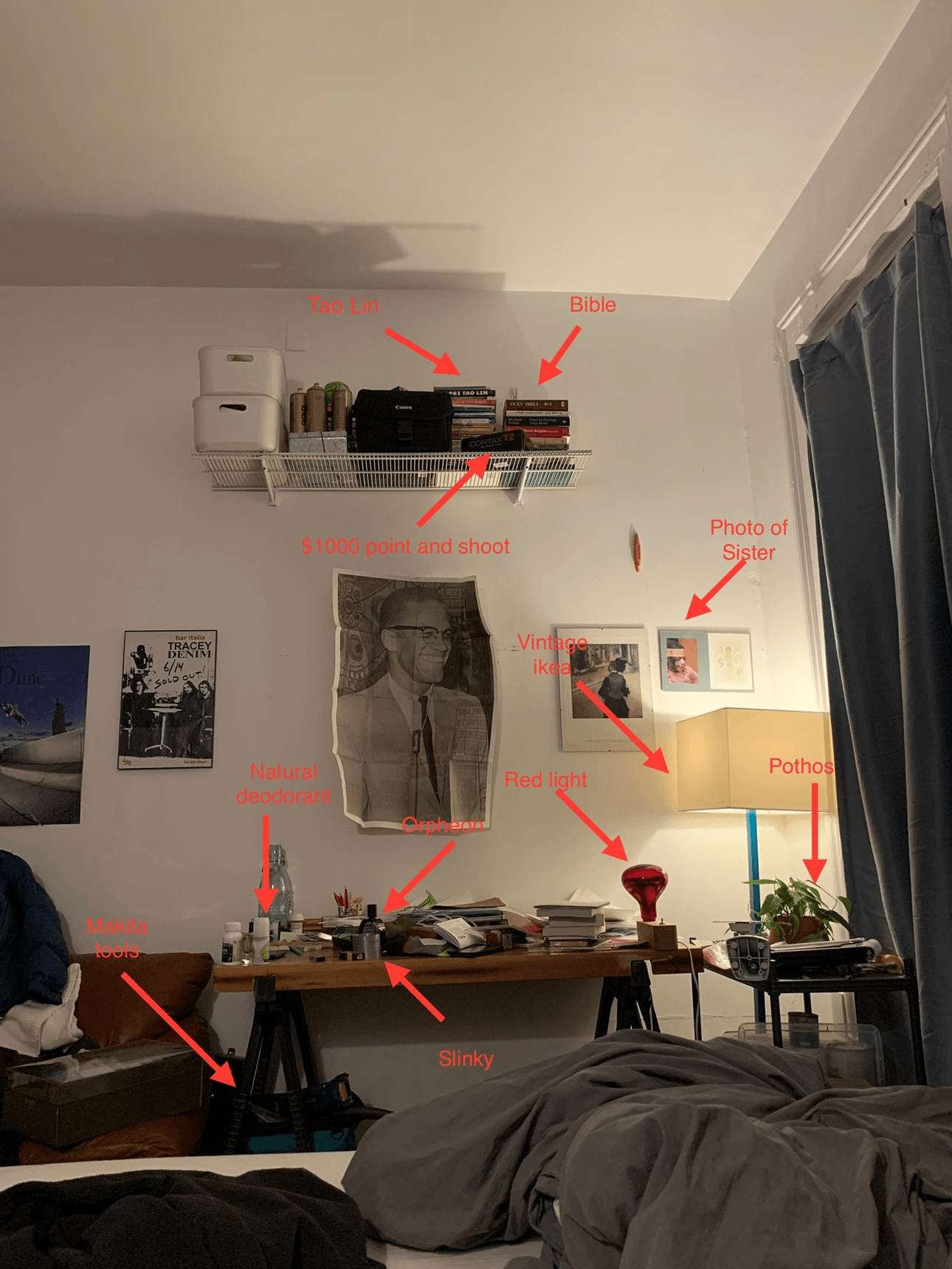 Bedroom scene at night showing a messy desk, lamp, wall art, plants, and labeled personal items like a Bible, photo, vintage camera, and toiletries; a bed with blankets is visible in the foreground.