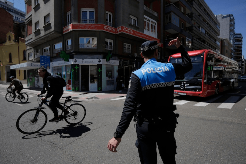 A police officer directs traffic at a city intersection as cyclists ride by and a red city bus waits at the crosswalk, with buildings and shops in the background.